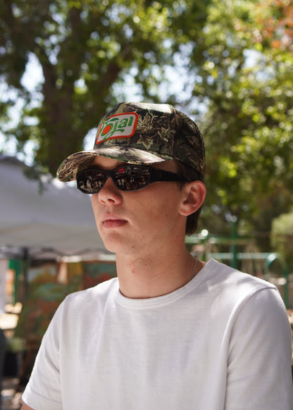 Person wearing a camouflage cap with a logo and sunglasses, standing outdoors with trees in the background.